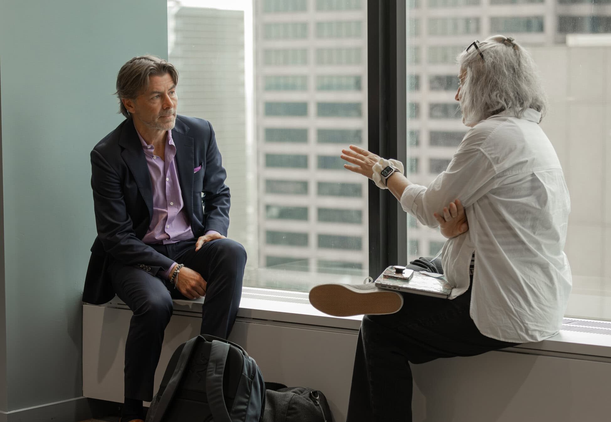 Casual Networking by the Window: Two professionals, a middle-aged Caucasian man in a suit and an older Caucasian woman with grey hair, having a serious discussion while sitting by a window overlooking the cityscape.